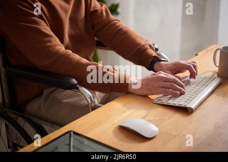 Closeup image of young man sitting in wheelchair working on laptop at his desk Stock Photo