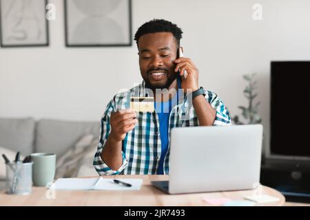 Cheerful Black Guy Holding Credit Card Talking On Phone Indoors Stock Photo