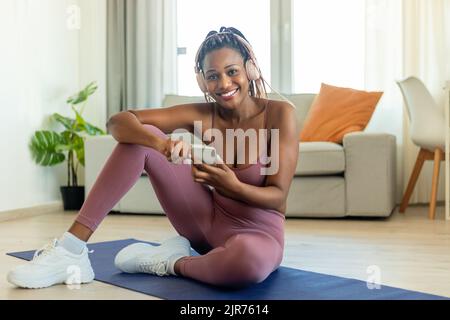 Smiling fit black woman sitting on yoga mat, using smartphone during workout break, listening music in headphones Stock Photo