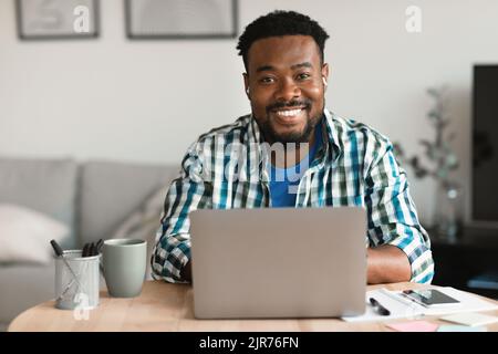 Cheerful Black Man At Laptop Computer Wearing Earbuds Working Indoors Stock Photo