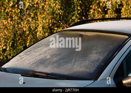 Frost on the windshield, yellow autumn leaves in the background. Autumn frosty morning. Frozen car window. Stock Photo