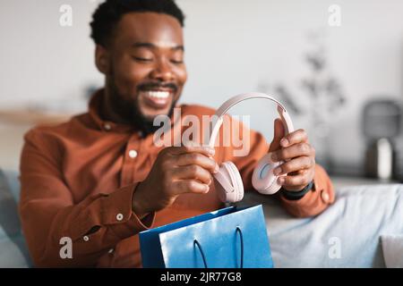 Happy African American Guy Unpacking Shopping Bag With Earphones Indoor Stock Photo