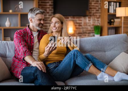 Happy middle aged caucasian wife show smartphone to husband, watching video, sitting on sofa Stock Photo
