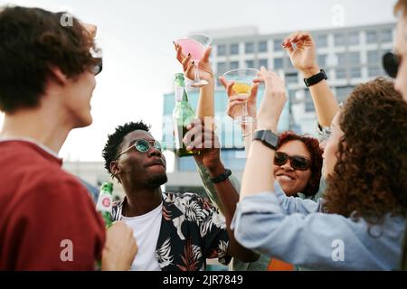 Group of cheerful friends raising hands with beer and cocktails while cheering up on terrace of rooftop cafe at outdoor party Stock Photo