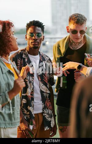 Group of intercultural young friends in casulawear having beer and chatting while standing in rooftop cafe during party Stock Photo