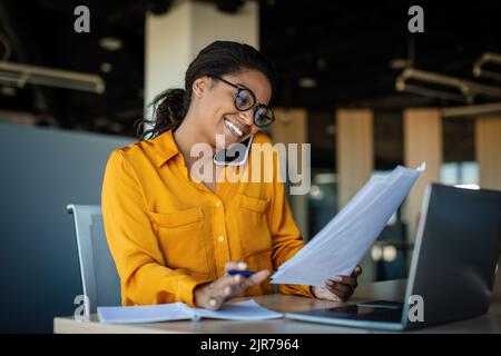 Corporate communication. Happy black female manager working with papers and talking on mobile phone Stock Photo