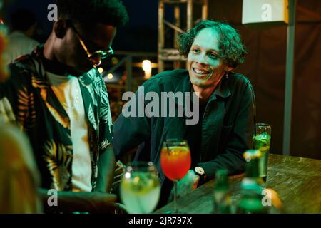 Happy young man in black casualwear looking at one of his friends during chat while sitting by counter with ordered cocktails in bar Stock Photo