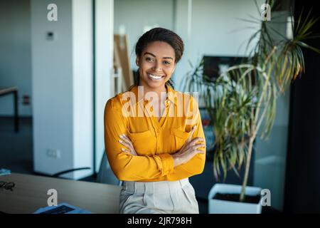 Happy black businesswoman standing with crossed arms in front of her desk at modern office and smiling at camera Stock Photo