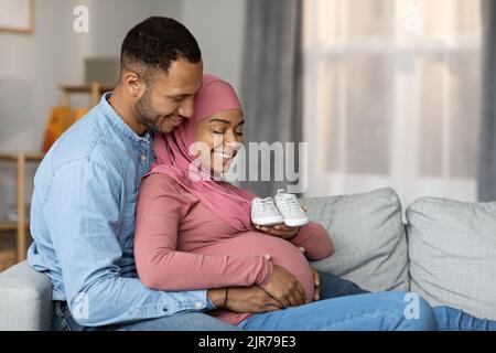 Parenthood Concept. Happy Black Muslim Couple Embracing And Holding Small Baby Shoes Stock Photo