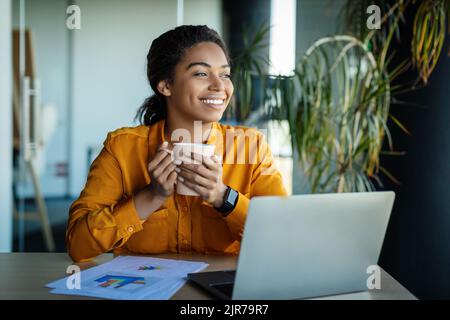Dreamy black businesswoman using laptop and holding coffee cup, looking aside and thinking, sitting at workplace Stock Photo