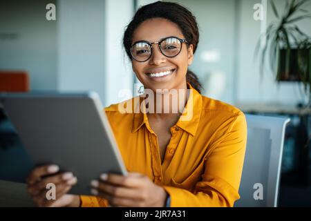 Happy african american business lady using tablet pc at workplace in office, looking and smiling at camera Stock Photo