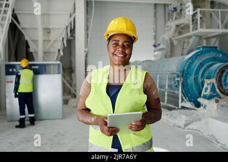 Happy young African American woman with tablet standing in front of camera in production plant or workshop and networking Stock Photo
