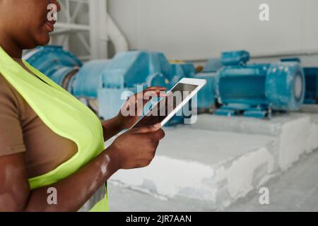 Hands of young African American female worker of factory using digital tablet while looking at sketch on screen of mobile gadget Stock Photo