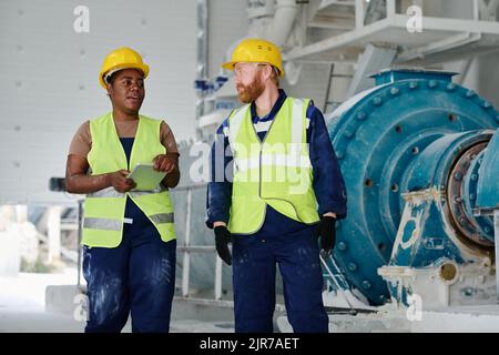 Two confident engineers in workwear and hardhats discussing working points at meeting while walking along factory workshop Stock Photo