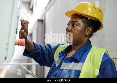 Young serious black woman in hardhat and workwear pressing button on panel while switching on production equipment in factory Stock Photo