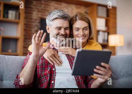 Cheerful mature caucasian female hugging man, waving hand, watching video in tablet, calling online Stock Photo