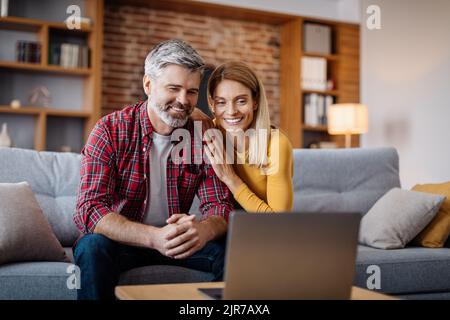Cheerful middle aged caucasian man and woman watch video in laptop, making online call in living room Stock Photo