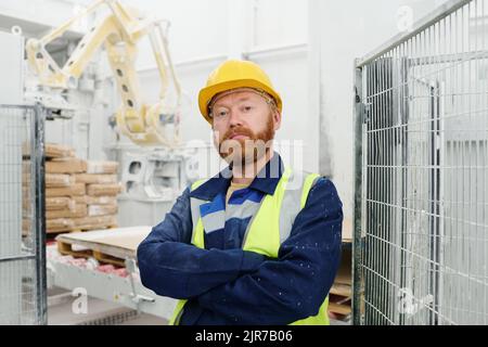 Confident bearded technician or engineer of factory looking at young while standing in front of camera in workshop or warehouse Stock Photo