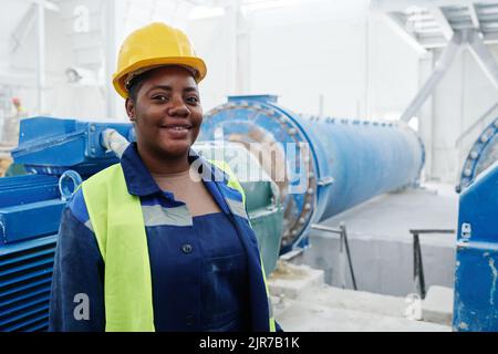 Happy young African American female engineer in hardhat and unidorm standing in workshop with industrial production equipment Stock Photo