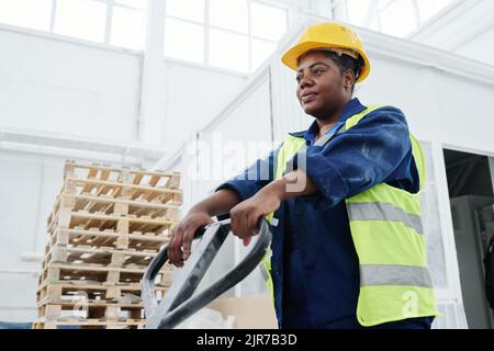 Young African American female worker of warehouse pushing forklift loader while moving along spacious workshop of factory Stock Photo