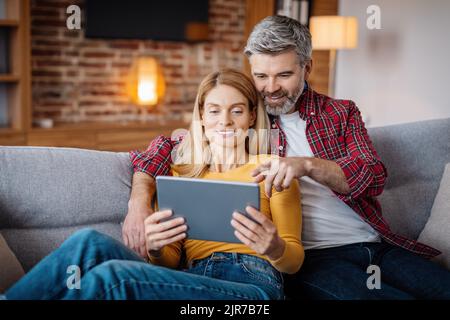 Smiling mature caucasian man hug lady and pointing finger at tablet, watch video in living room interior Stock Photo
