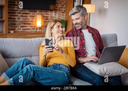 Glad adult caucasian female show phone to man with laptop, watch video in living room interior Stock Photo