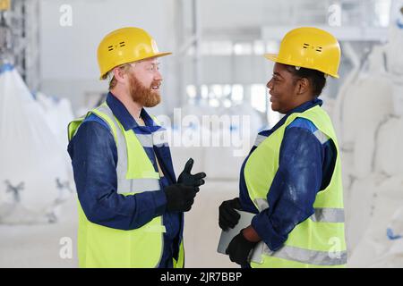 Side view of two young intercultural workers of factory in uniform and hardhats discussing characteristics of new machinery equipment Stock Photo