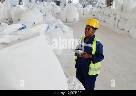 Young serious black woman in uniform and safety helmet using tablet while standing in front of huge white sack with raw materials Stock Photo