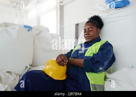 Tired female engineer in uniform holding safety helmet while sitting against huge white sack in warehouse and napping Stock Photo