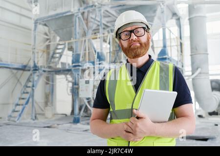 Confident bearded foreman in uniform and safety helmet holding tablet while standing in workshop of modern production factory Stock Photo