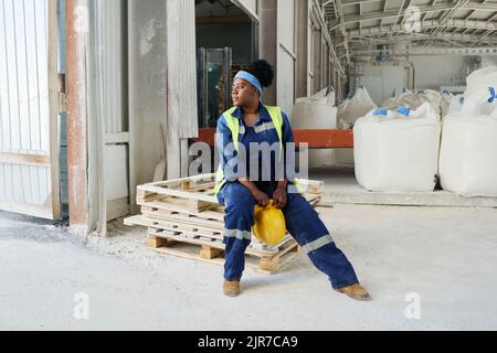 Young distracted African American female engineer with safety helmet in hands sitting on stack of wooden pallets in warehouse Stock Photo