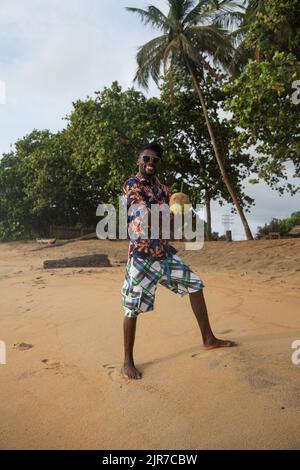 Barefoot on the sand, young man in sunglasses smiles and holds a coconut ready to be tasted. Stock Photo