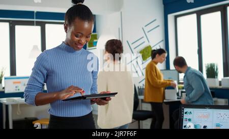 Portrait of african american woman standing in busy office picking up tablet with business erp software and smiling looking at touchscreen. Employee holding digital device and interacting with screen. Stock Photo