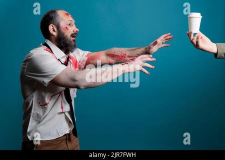 Scary wounded corpse wanting coffee cup in studio over blue background, going after desired drink and acting aggressive. Cruel undead zombie with bloody scars wishing for beverage. Stock Photo