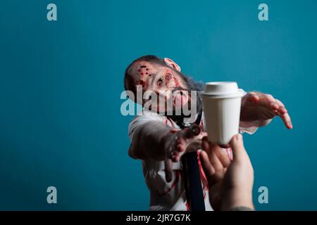 Aggressive halloween corpse wishing to drink coffee cup in studio, going after beverage and wanting to eat carbon. Spooky undead scary devil chasing hand with drink, acting dangerous and creepy. Stock Photo