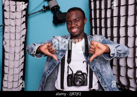 African american studio producer showing thumbs down hands symbol while having DSLR camera and professional photo equipment. Smiling photographer gesturing disapproval sign on blue background. Stock Photo