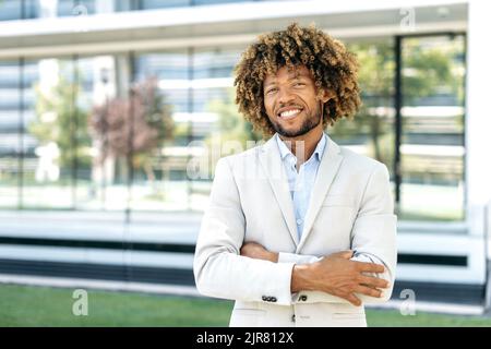 Elegant portrait of a happy confident successful curly haired brazilian or hispanic man, standing outdoors with arms crossed near business center, looking at the camera, smiling friendly Stock Photo