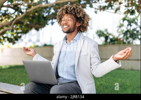 Positive calm curly haired brazilian or hispanic man, company employee, entrepreneur, sits on outdoors with laptop in his hands, takes pause at work, meditation with eyes closed, relaxing, smiles Stock Photo
