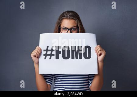 If its happening I want to be part of it. Studio shot of an attractive young woman holding a sign with FOMO printed on it against a gray background. Stock Photo