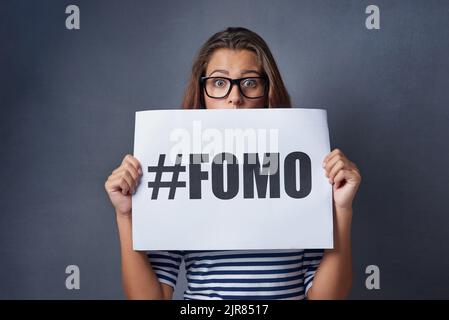 If it happened online it must be real. Studio shot of an attractive young woman holding a sign with FOMO printed on it against a gray background. Stock Photo
