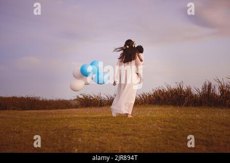 They share a close bond. Rearview shot of a young mother holding her son and balloons outdoors. Stock Photo