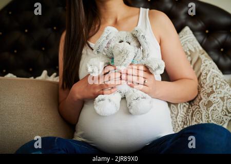 Playtime starts before her due date. an unidentifiable pregnant woman holding a stuffed toy while sitting on her bed at home. Stock Photo