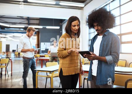 Let me check it out. two young businesspeople looking at a tablet in the office with their colleagues in the background. Stock Photo