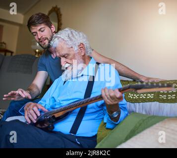 Teaching him a riff or two. a happy young man teaching his elderly grandfather to play the electric guitar on the couch at home. Stock Photo
