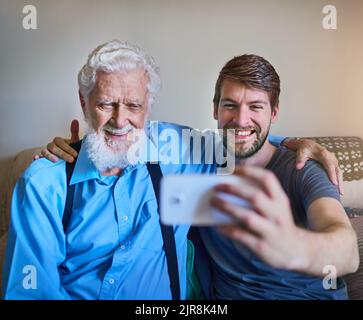 These are the moments we want to cherish. a happy young man and his elderly grandfather posing for a selfie together on the couch at home. Stock Photo