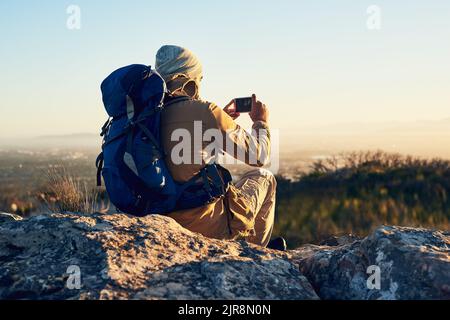 It takes time to capture the perfect picture. a hiker on top of a mountain taking a photo with his cellphone. Stock Photo