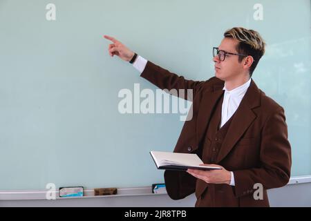 A teacher pointing at a blackboard. University professor with a black book in his hand. Master dressed in an elegant brown suit. Elegant man in a blackboard explaining a topic. High quality photo Stock Photo