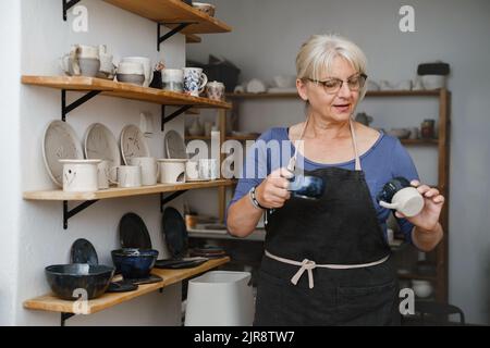 Potter artist showing her work in studio. Mature ceramist standing at workshop with clay cups in hands. Successful lady potter own making porcelain Stock Photo