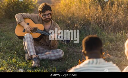 A young bearded caucasian man playing the guitar during a picnic in nature with his friends. People chilling near the fire, enjoying and watching the sunset. High quality photo Stock Photo