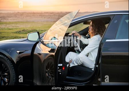 Side view of cheerful female owner of agribusiness sitting in black car and smiling while thinking about land tenure in nature environment, happy Caucaisan woman in automobile enjoying lifestyle Stock Photo